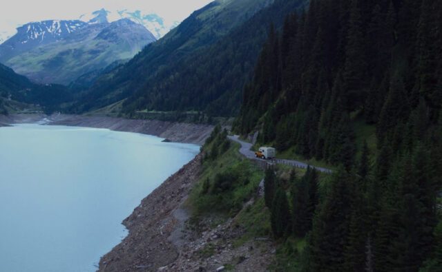 tischer_mieten_Tischer Wohnkabine auf einem Pick-up unterwegs auf einer kurvigen Uferstraße inmitten alpiner Berglandschaft – unterwegs in freier Natur.